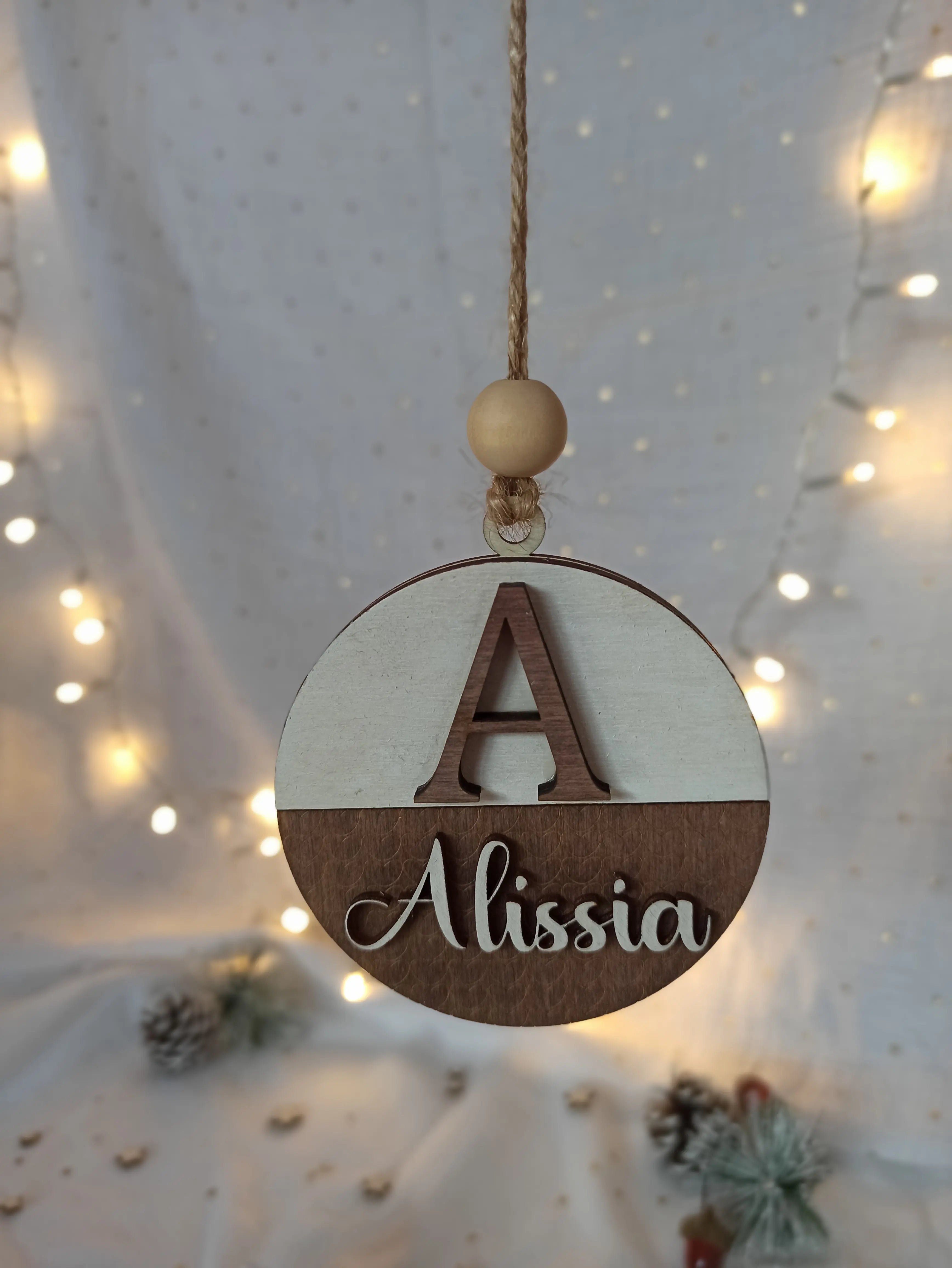 Suspension de Noël personnalisée avec prénom et initiale en bois, suspendue devant des guirlandes lumineuses pour une ambiance chaleureuse et poétique.